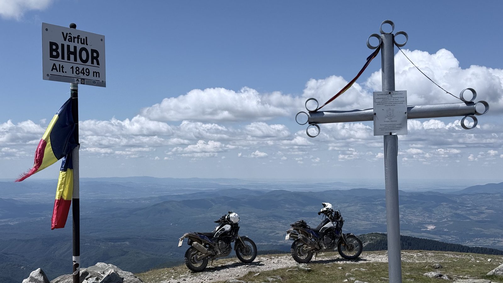 Two Yamaha T7 motorcycles on the summit of Vârful Bihor at 1,849 m, beside the summit sign, a Romanian flag and a metal cross
